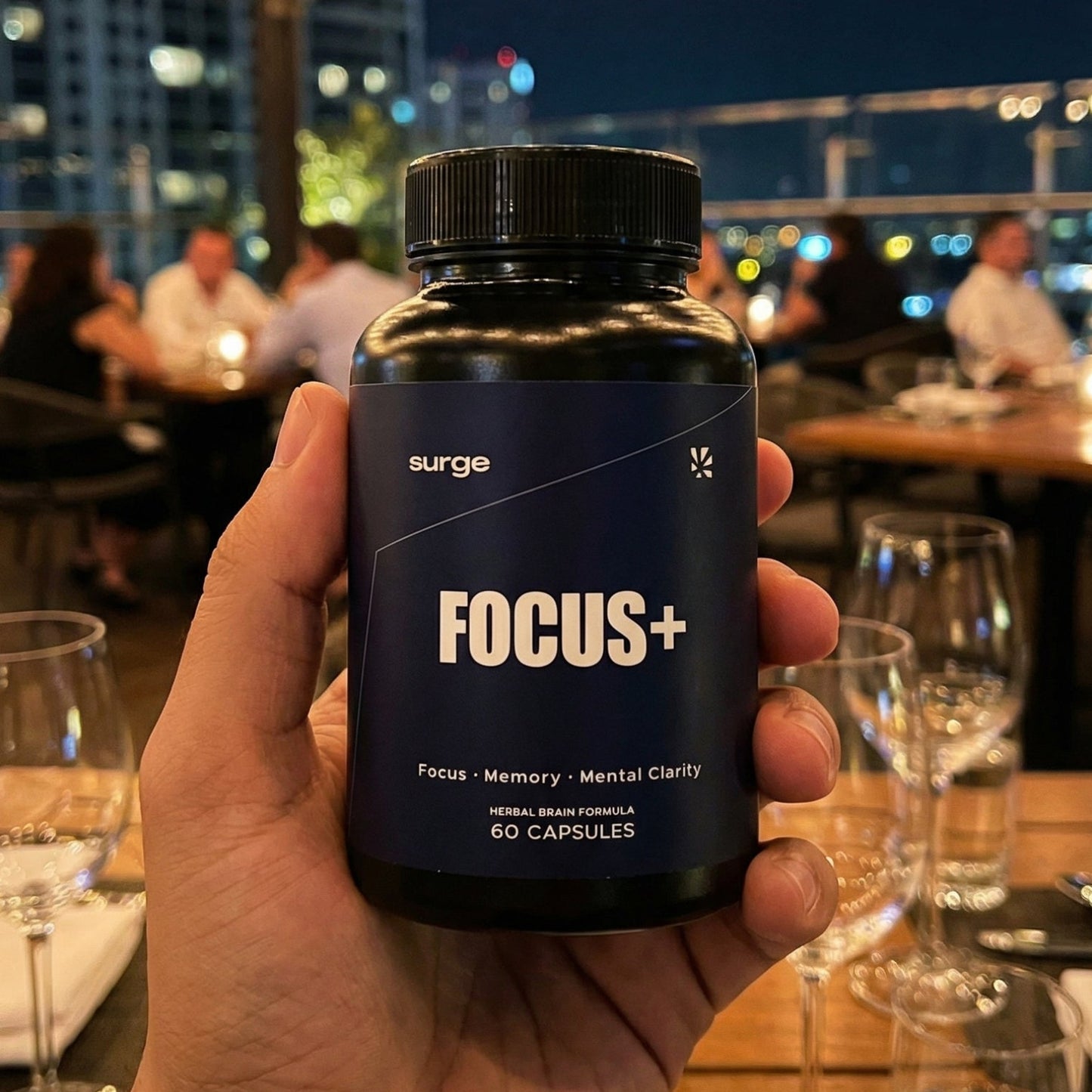 Hand holding a bottle labeled 'FOCUS+' in an outdoor evening setting with lights and people in the background.